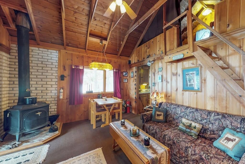 A rustic cabin interior with a brick wall, wooden furniture, a sofa with animal-themed cushions, a table with decorative items, and a staircase.