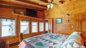 Ein Schlafzimmer einer Holzhütte mit Bett, Fenstern, Deckenventilator und einer gesteppten Decke.