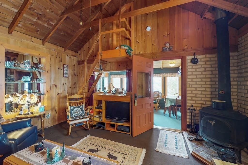 Interior rústico de una cabaña con escalera de madera, televisor, chimenea y comedor.