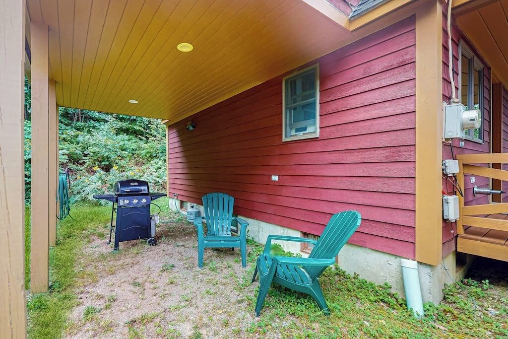 Un espace extérieur abrité, équipé d’un barbecue, de deux fauteuils Adirondack verts et d’une clôture en bois.