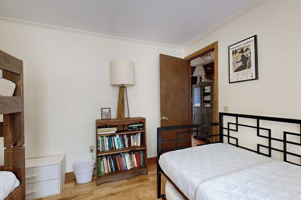 A bedroom with a bed, a wooden bookshelf, a white lamp, and a door leading to a bathroom.