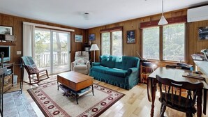 A spacious living room with a wooden floor, a blue sofa, a wooden coffee table, a dining table with chairs, and large windows offering a view of the outdoors. - Lady Slipper Cottage: Charming Lakefront Retreat With Canoes, Kayaks, and Modern Comforts (Mount Desert)