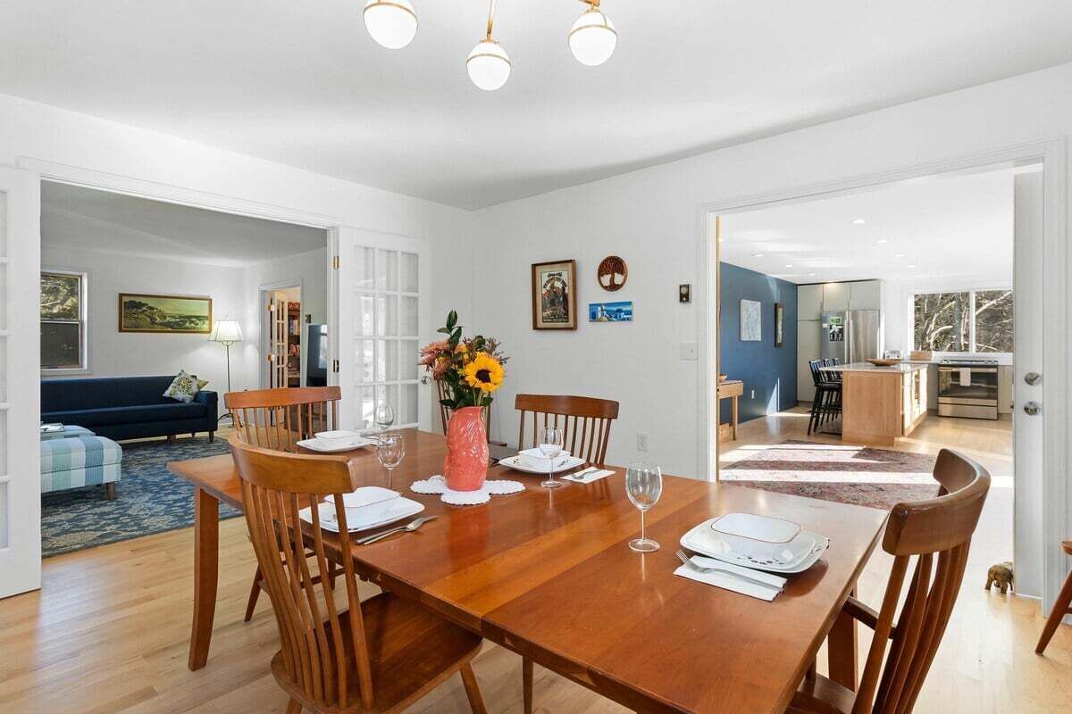 A dining area with a wooden table set for four, a vase with flowers, and a view into the kitchen.