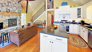 A spacious living area with a stone fireplace, a brown sofa, a kitchen with white cabinets, and a wooden staircase. - Renovated Waterfront Cottage With Expansive Deck, Multiple Fireplaces, & Grill (Ellsworth)