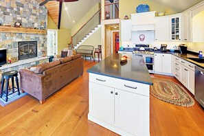 Ein großzügiger Wohnbereich mit Kamin aus Stein, einem braunen Sofa, einer Küche mit weißen Schränken und einer Holztreppe.