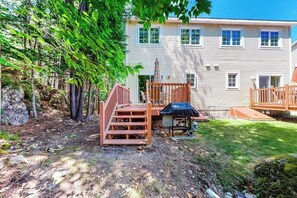 A two-story house with a deck, a covered grill area, and a stone wall. - Convenient Home With Deck, Grill, & Washer/dryer - Walk Into Town & Acadia (Bar Harbor)