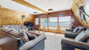 A log cabin interior with a sofa, a television, and a view of the outdoors. - Comfy Lakefront Cabin With Gorgeous Views, Private Dock & Pebble Beach (Greenville)
