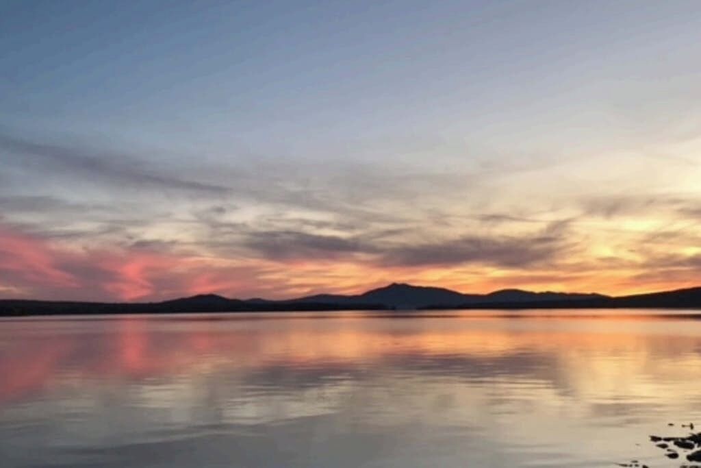 A calm lake with a sunset, mountains in the distance, and a clear sky.