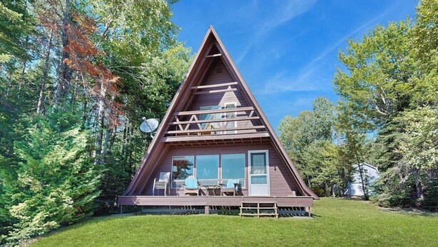 A triangular wooden cabin with a deck, large windows, and a dining area. - Comfy Lakefront Cabin With Gorgeous Views, Private Dock & Pebble Beach (Greenville)