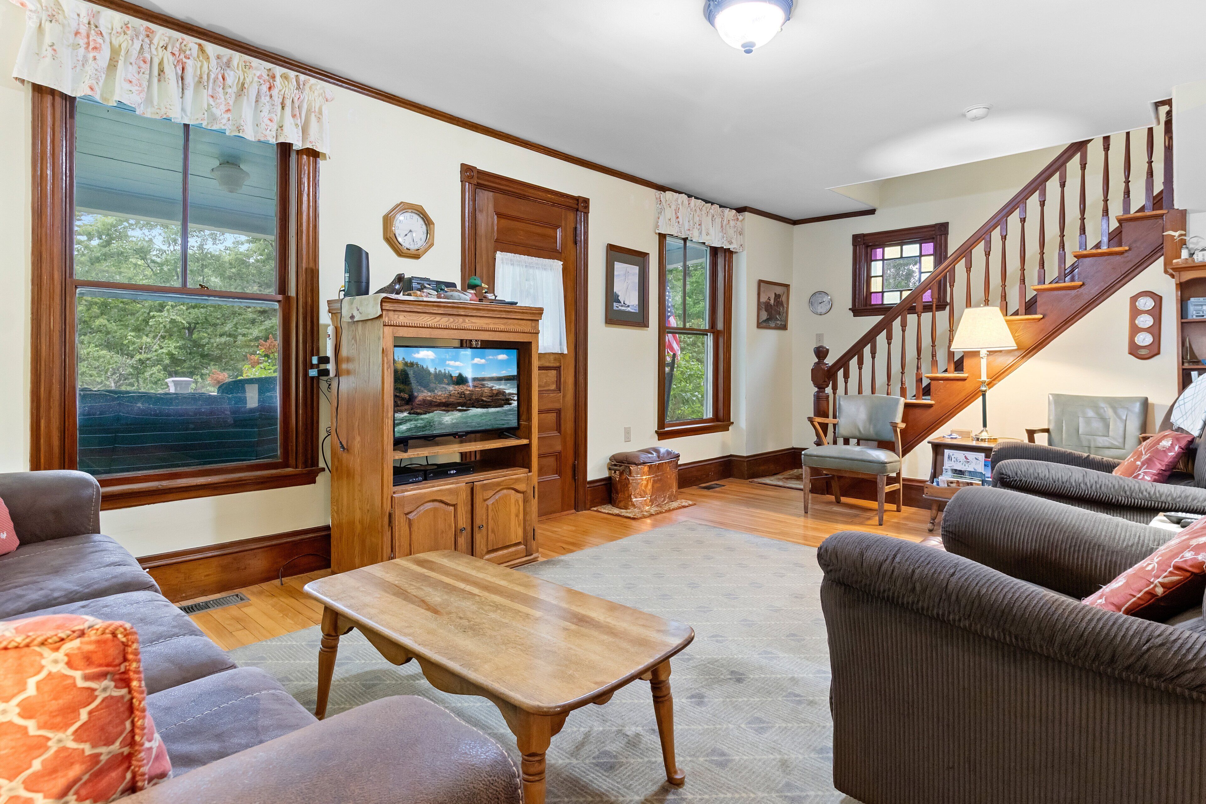 A living room with a television, wooden furniture, a staircase, and large windows.