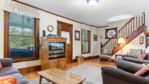 A living room with a television, wooden furniture, a staircase, and large windows. - Century-old Oceanfront Home With Wraparound Porch - Walk Downtown, Dogs OK (Boothbay Harbor)