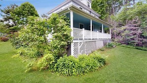 A two-story house with a covered porch, surrounded by lush greenery and trees. - Century-old Oceanfront Home With Wraparound Porch - Walk Downtown, Dogs OK (Boothbay Harbor)