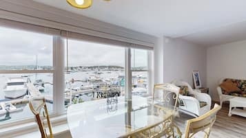 A modern living room with a glass dining table, white wicker furniture, and large windows overlooking a marina.