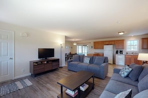 A living room with a sofa, a coffee table, and a TV. There is a kitchen in the background with wooden cabinets and a refrigerator. - Tranquil Getaway on the Coast With Beach Access, Ocean Views, & Screened Porch (Tenants Harbor)