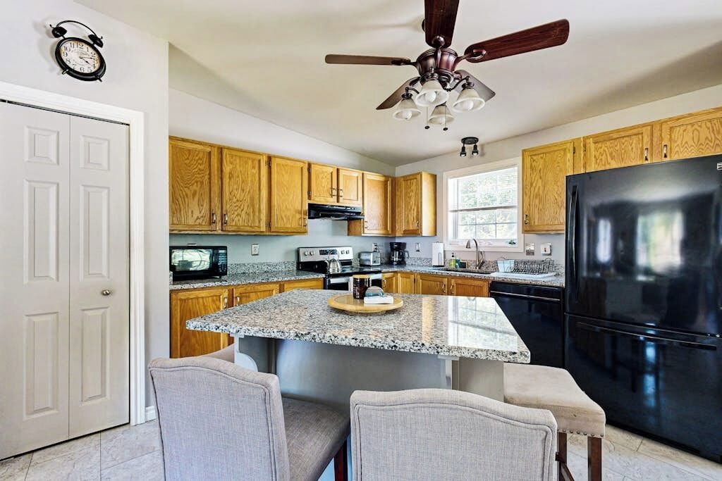 A kitchen with wooden cabinets, a black refrigerator, a granite countertop island, and a ceiling fan.