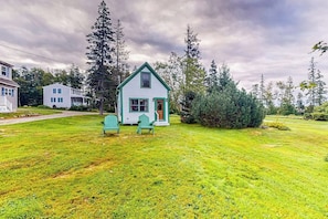 A small white house with a green roof, surrounded by a grassy yard and tall trees. - Well-equipped Cottage With Modern Touches, Wifi, gas Grill, & Beautiful Views (Port Clyde)