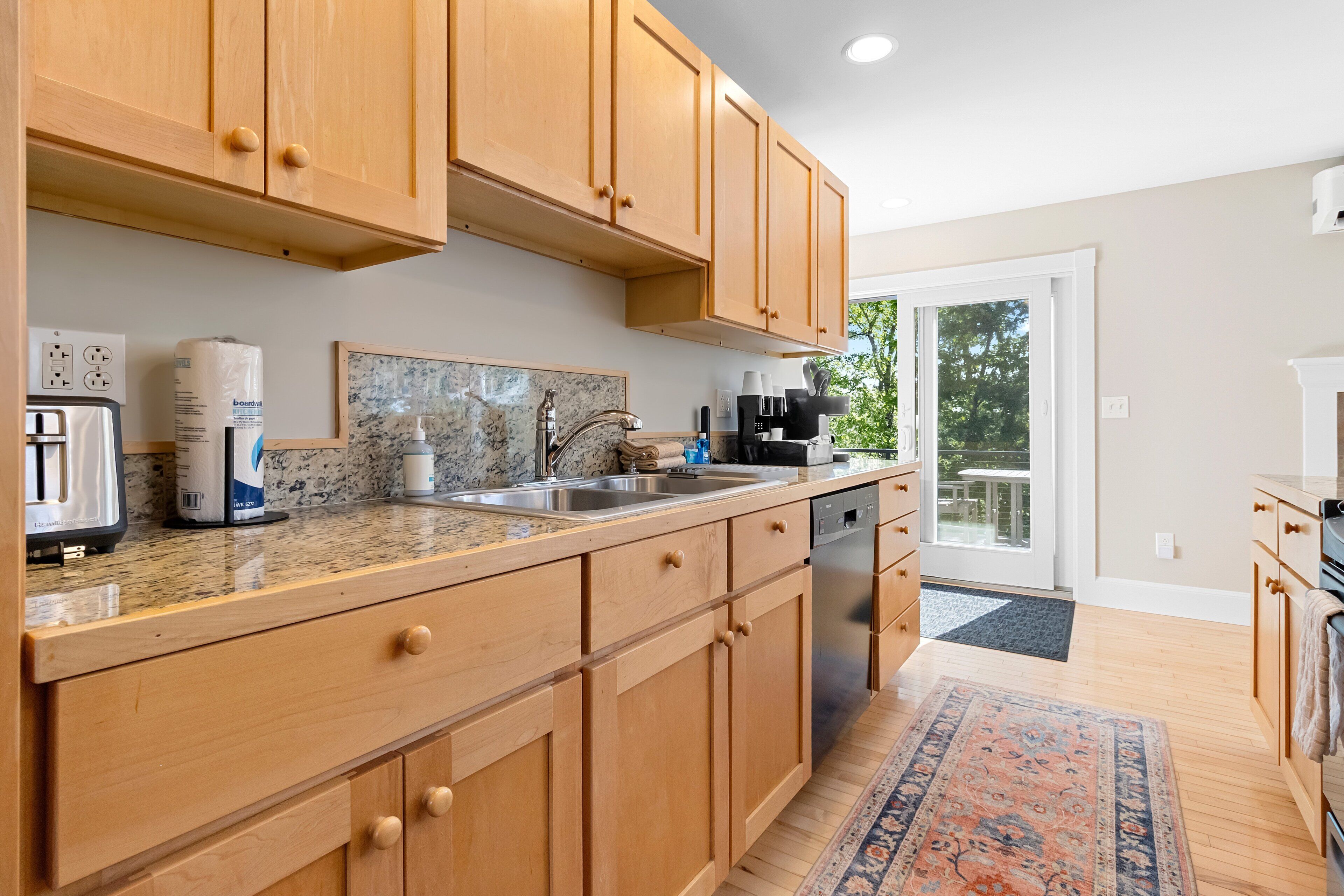 A kitchen with wooden cabinets, a granite countertop, a built-in dishwasher, and a view to the outside.