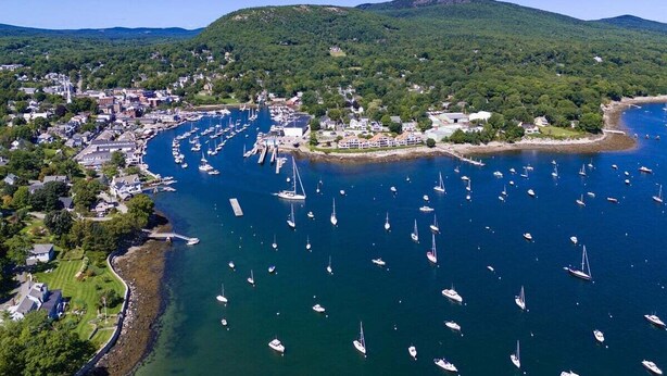 An aerial view of a coastal town with a marina filled with boats, surrounded by green hills and residential buildings. - Historic Condo in Downtown Camden, Steps to Harbor (Camden)