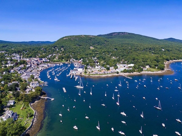 An aerial view of a coastal town with a marina filled with boats, surrounded by green hills and residential buildings. - Historic Condo in Downtown Camden, Steps to Harbor (Camden)
