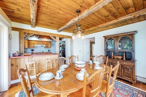 A wooden dining table set with plates, cups, and cutlery, surrounded by chairs, in a room with exposed wooden beams and a large window. - Dog-friendly, Bayfront Home W/rocky Beach, Full Kitchen, Washer/dryer & Firepit (Gouldsboro)