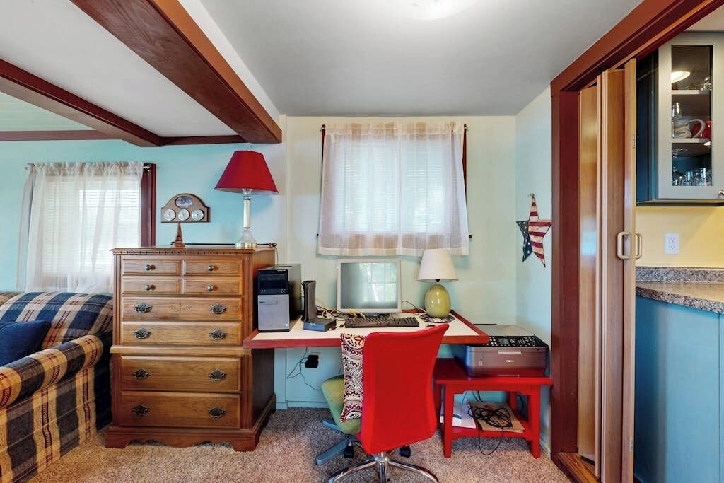 Una habitación con un escritorio de madera, una computadora, una silla roja, un aparador y una ventana con cortinas.