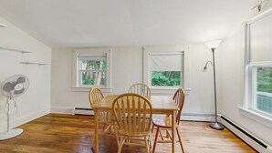 Un comedor con mesa y sillas, un ventilador de pie y ventanas con vistas a zonas verdes.