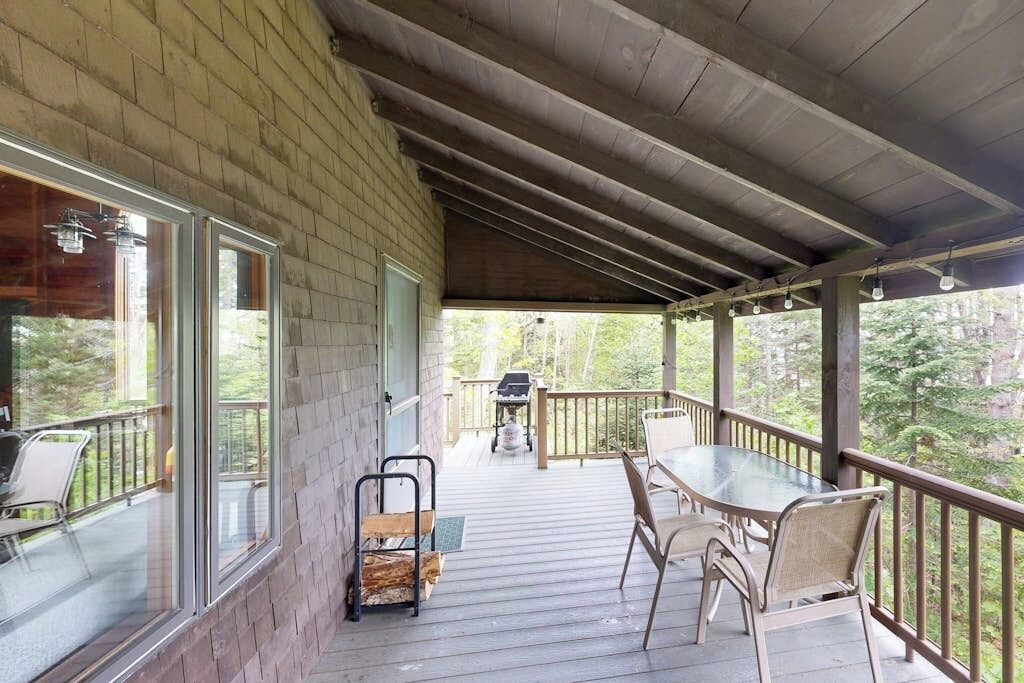 A covered outdoor deck with a table and chairs, a grill, and a view of the surrounding forest.