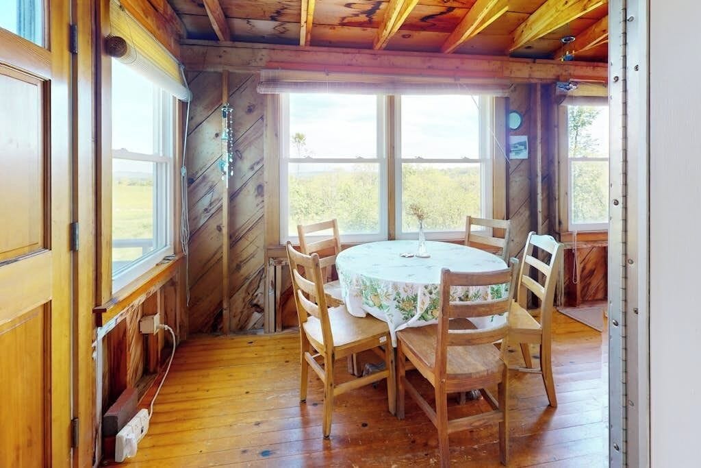 A wooden dining room with a round table and six chairs, large windows, and a view of the outdoors.
