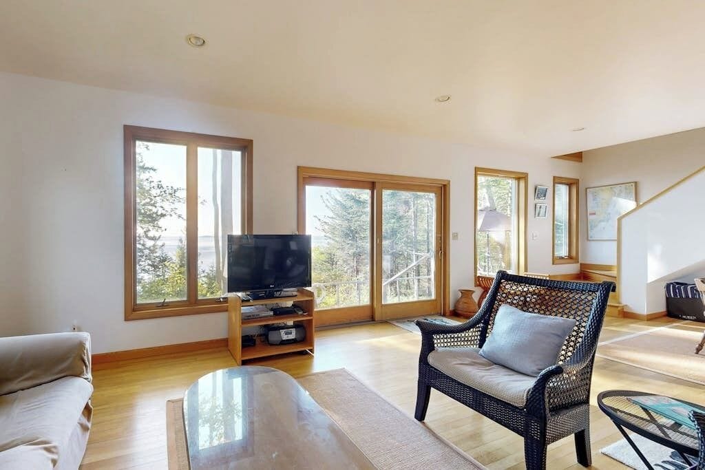 A spacious living room with a television, a sofa, a wicker chair, and large windows.