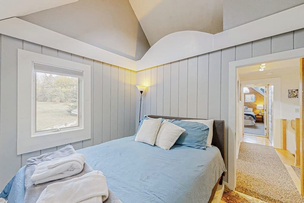 A bedroom with a bed, a window, a lamp, and a door leading to another room.