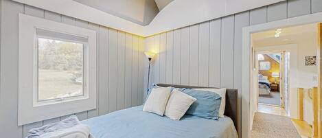 A bedroom with a bed, a window, a lamp, and a door leading to another room.