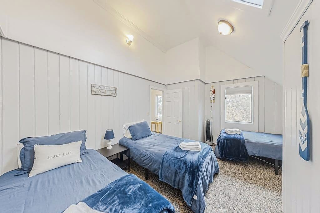 Ein Zimmer mit drei Betten, einem kleinen Tisch, einer Lampe und einem Fenster.
