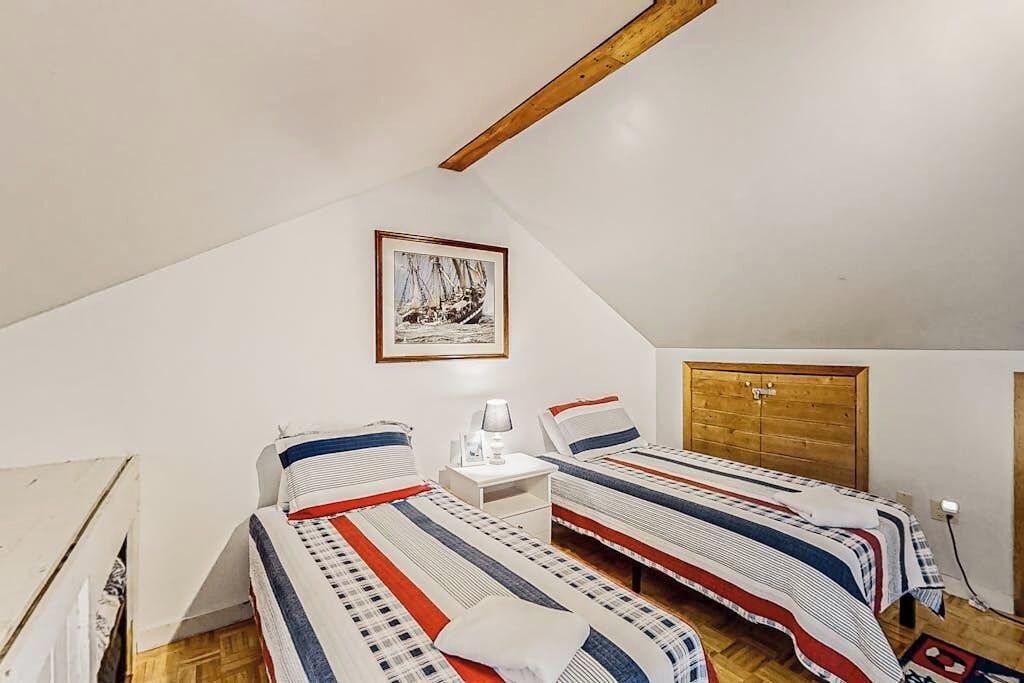 A room with two beds, a wooden ceiling beam, a framed picture on the wall, and a white bedside table.