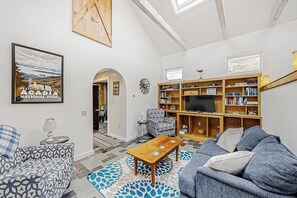Ein Wohnzimmer mit einer Couch, einem Couchtisch, einem Bücherregal, einem Fernseher und einem gerahmten Bild an der Wand.
