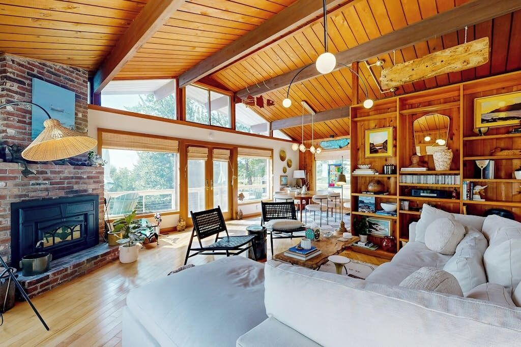 A spacious living room with a fireplace, wooden ceiling, and large windows.