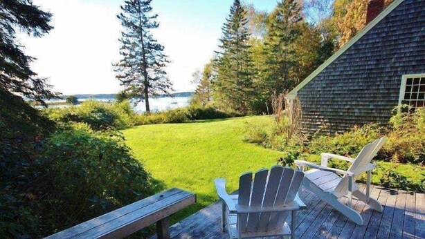 A deck with Adirondack chairs and a bench, overlooking a grassy yard with a house and trees. - Tideriver, Waterfront Cottage in Maine (Port Clyde)