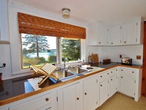 A kitchen with white cabinets, a wooden countertop, a window with a view of trees and a body of water, and a built-in sink. - Tideriver, Waterfront Cottage in Maine (Port Clyde)