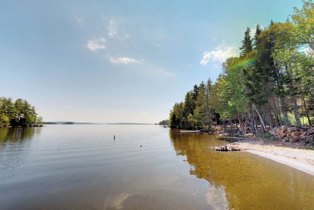 A calm lake with trees on both sides, a clear sky, and a rainbow.