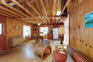 Interior de cabana com paredes de madeira, área de refeições com cadeiras, cozinha com geladeira e uma janela.