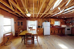 Interior de uma cabana de madeira com mesa de jantar, cadeiras, cozinha equipada e uma janela.