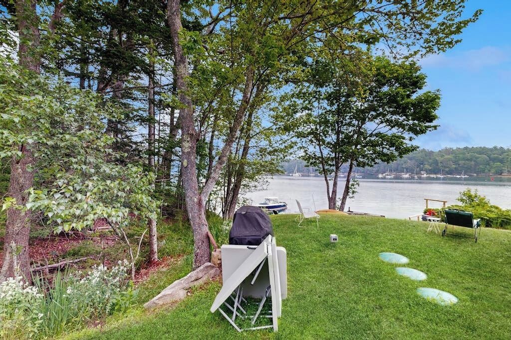 Un espacio al aire libre con césped, una silla plegable blanca, una parrilla y vistas a una masa de agua con barcos.