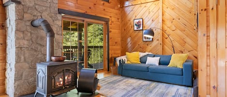 A cozy cabin interior with a stone wall, a fireplace, a blue sofa with cushions, and a large window.