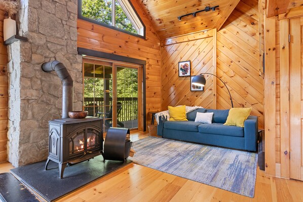 Un interior de cabaña acogedor con pared de piedra, chimenea, sofá azul con cojines y un ventanal grande.