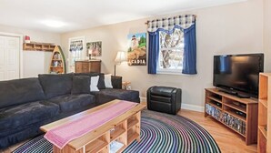 A living room with a sofa, a coffee table, a TV, a bookshelf, and a window with curtains. - Dog-friendly Home in Great Locale Close to the Ocean With Yard, Streaming, W/D (Seal Cove)