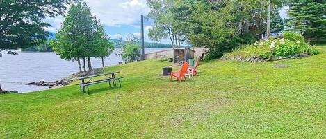 Ein Rasen mit Picknicktisch und -stühlen, ein kleines Gartenhaus und Bäume in der Nähe eines Gewässers.