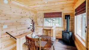 A wooden cabin interior with a dining table, chairs, a vase with flowers, a window, and a wood-burning stove. - Charming Riverfront Home With Mountain Views & Private Dock (Rockwood)