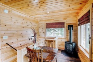 Interior de uma cabana de madeira com mesa de jantar, cadeiras, um vaso com flores, uma janela e uma lareira a lenha.