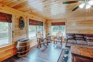 Interior de uma cabana de madeira com sofá, poltronas de balanço, mesa e um barril.