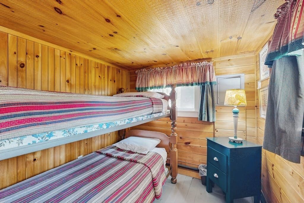 Une chambre de style cabane avec deux lits superposés, un plafond en bois, une petite table de chevet avec une lampe et une fenêtre avec des rideaux.