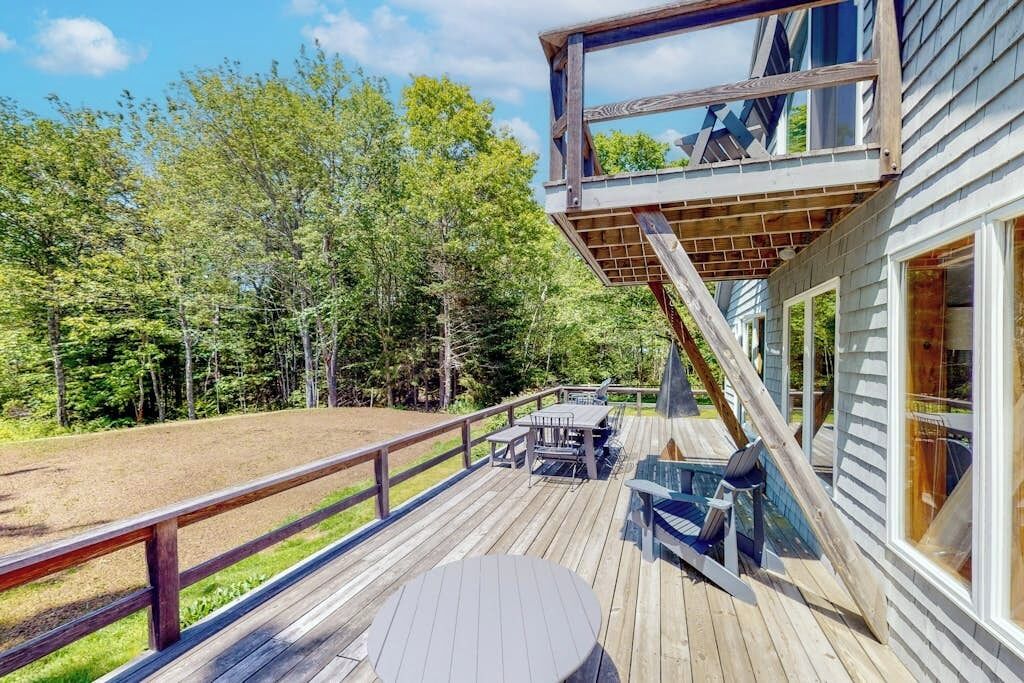 A wooden deck with outdoor furniture, a staircase leading to a second-story deck, and a view of a forested area.